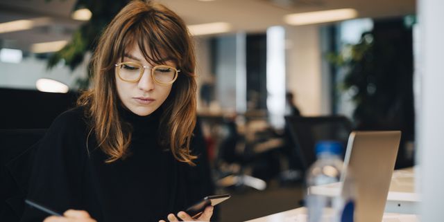 Businesswoman writing while holding mobile phone at desk in office