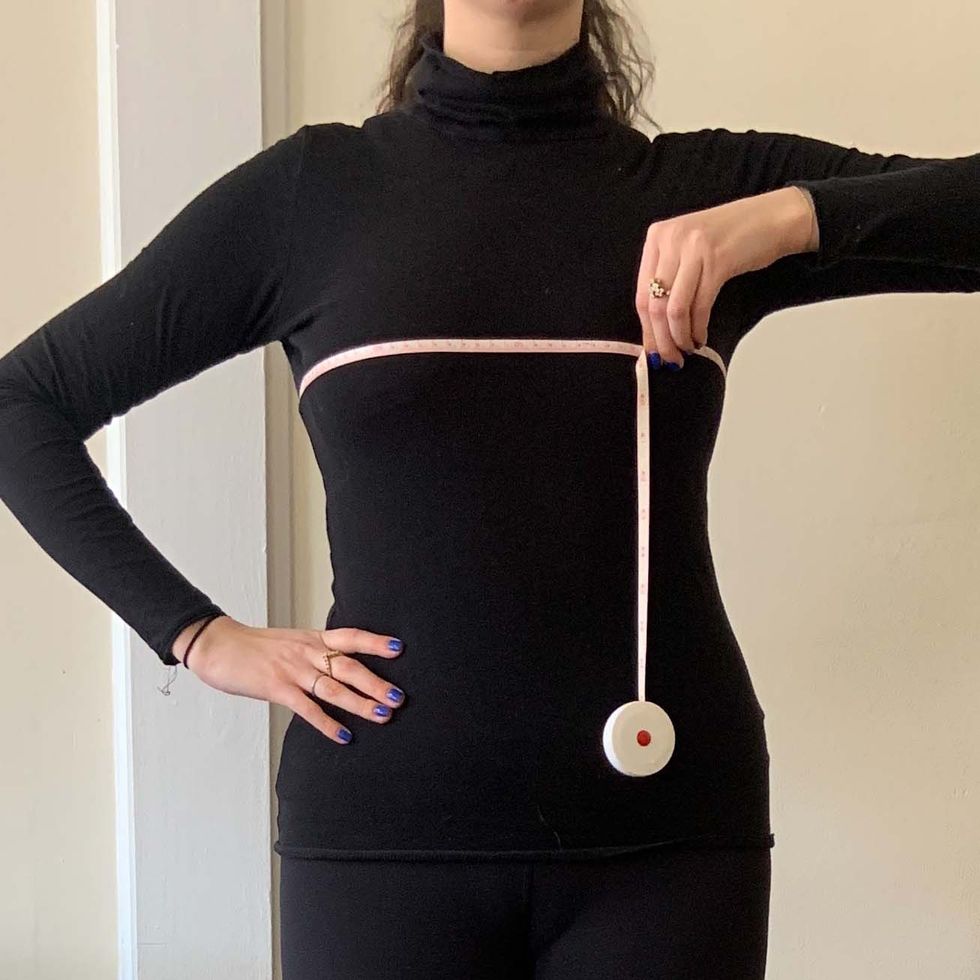 a woman wearing black leggings and a black turtle neck demonstrate how to measure your bust with a measuring tape straight across her chest