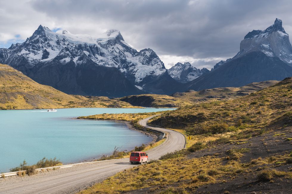 a red car driving on a road by a lake and mountains