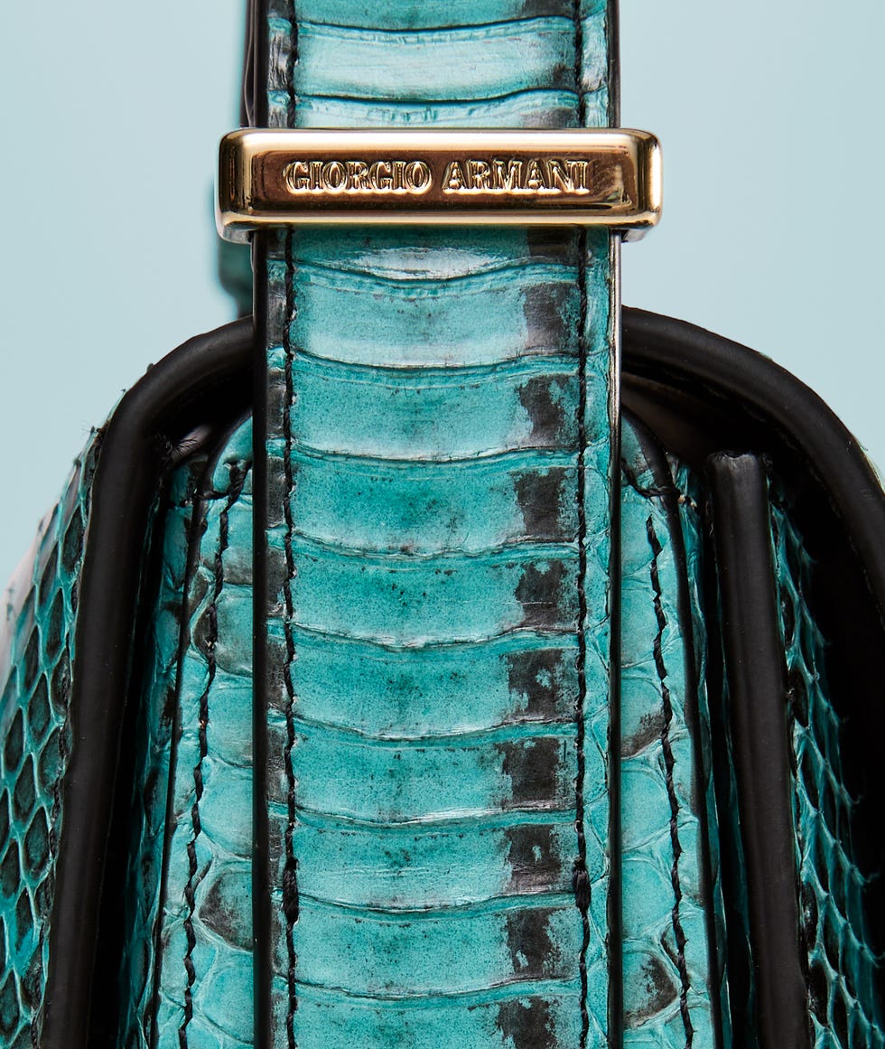 Close-up of a handbag strap featuring bold turquoise crocodile texture and a gold logo plaque.