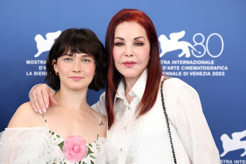 cailee spaeny and priscilla presley pose for a photo while standing in front of a blue background, spaeny wears a floral spaghetti strap dress with a pink flower in the center and tulle sleeves, presley wears a white bejeweled collared shirt and has one arm around spaenys shoulders