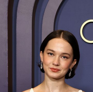 cailee spaeny smiles at the camera, she wears a cream colored dress and dangling diamond earrings and stands in front of a navy blue background