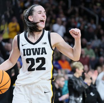 caitlin clark holds a basketball in one hand and pumps her other fist, she wears a white and black basketball uniform for iowa and smiles
