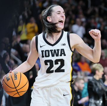 caitlin clark holds a basketball in one hand and pumps her other fist, she wears a white and black basketball uniform for iowa and smiles
