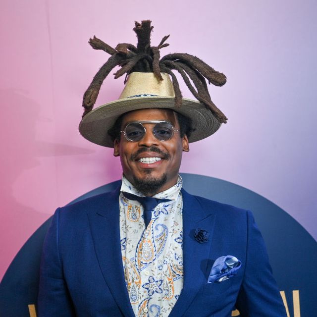 cam newton smiles at the camera, he wears a royal blue suit jacket and matching tie, colorful paisley shirt, blue tinted glasses, and straw hat with his dreadlocks falling over the front of the hat