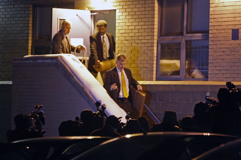 three men in suits and ties walk out of a building at night and down steps as photographers and videographers wait in a dark crowd in the foreground, each man is holding items in their hands