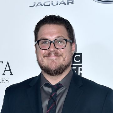 cameron britton, a man with short brown hair, a short beard and black glasses, wearing a blue suit with grey shirt and tie, smiling at the camera