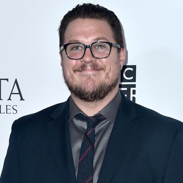 cameron britton, a man with short brown hair, a short beard and black glasses, wearing a blue suit with grey shirt and tie, smiling at the camera