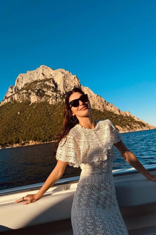 mulher em vestido de crochê branco posando em um barco com uma montanha ao fundo
