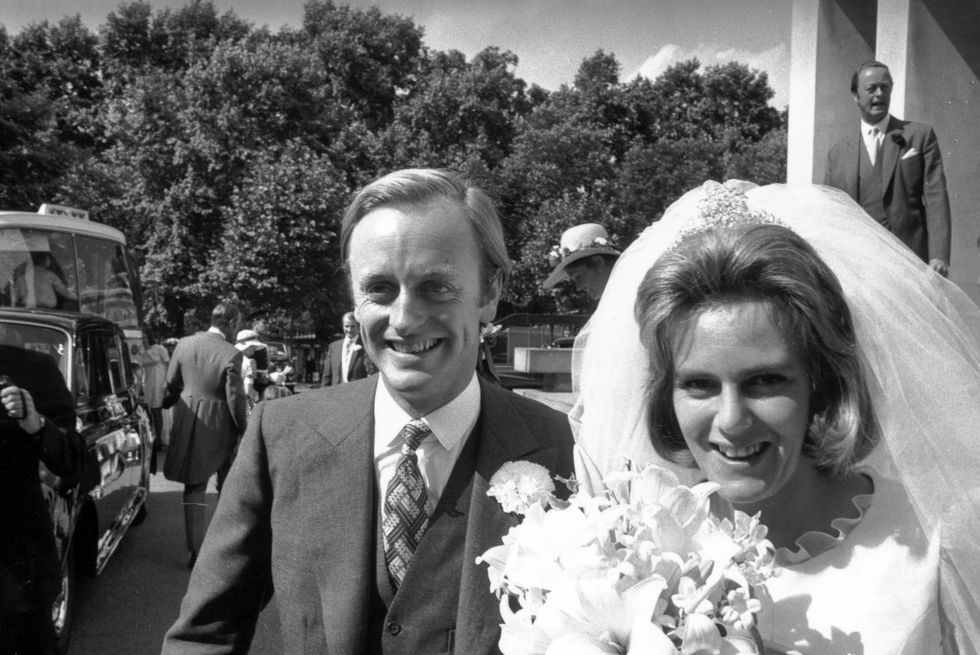 andrew parker bowles and camilla parker bowles stand outside a building arm in arm, he wears a suit with a boutonniere, she wears a white dress and veil and holds a bouquet