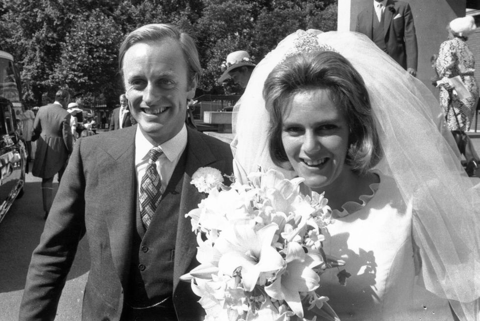andrew parker bowles and camilla parker bowles stand outside a building arm in arm, he wears a suit with a boutonniere, she wears a white dress and veil and holds a bouquet