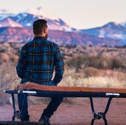 man wearing plaid flannel shirt sitting on camping cot by fire in desert looking at mountains