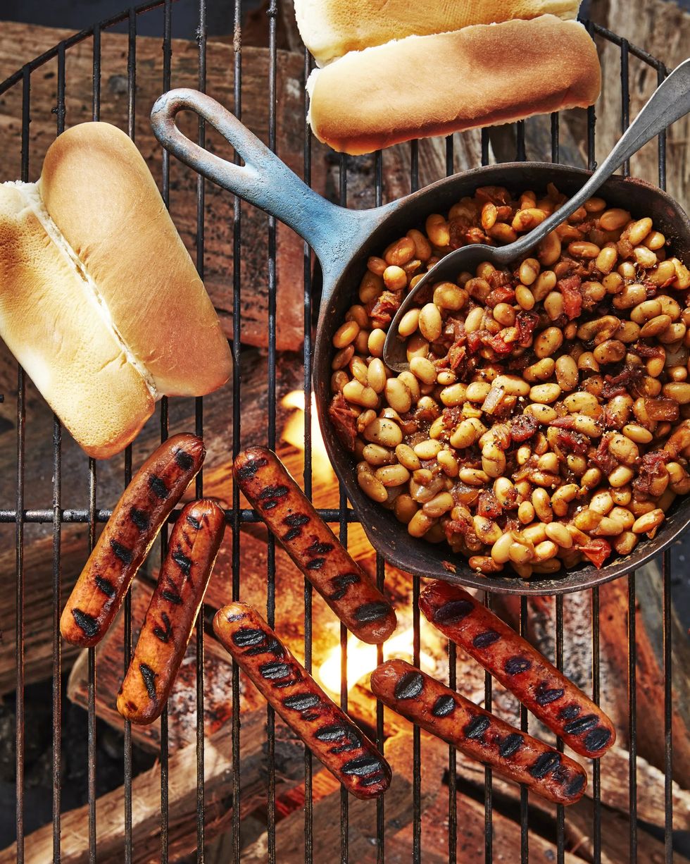 grilled hot dogs and buns on a grate over a campfire with a cast iron skillet of beans next to them