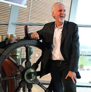 james cameron wearing a black suit jacket and white shit, smiling and leaning against a ship steering wheel