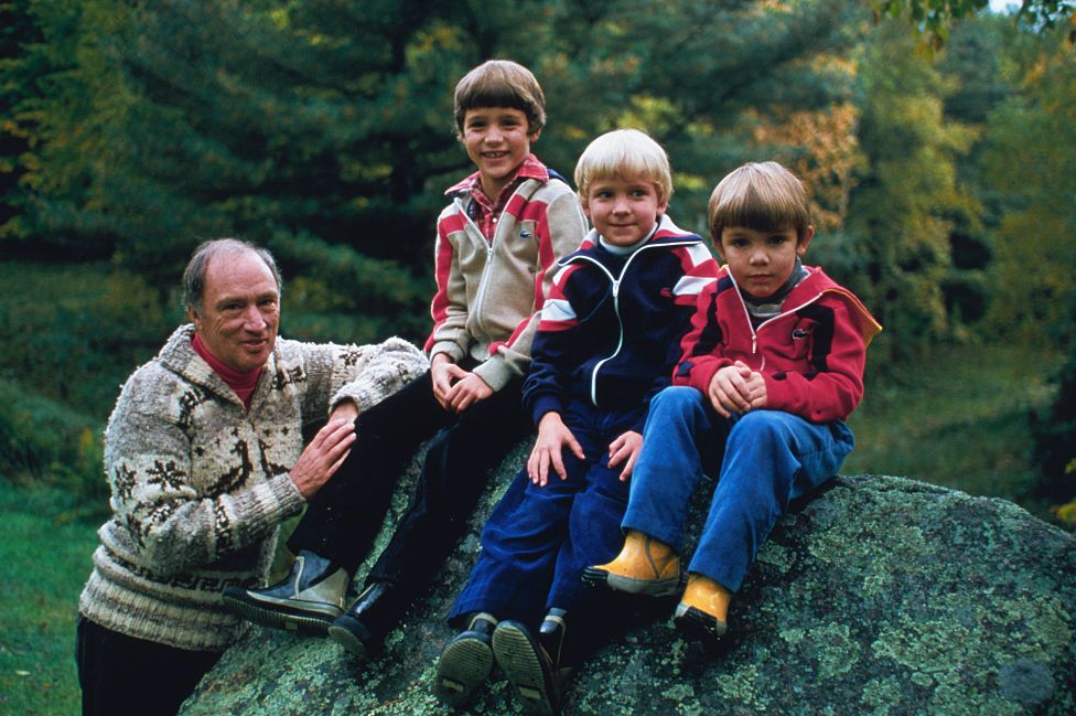 pierre trudeau smiles while leaning against a large boulder, his sons justin, alexandre and michel sit on the boulder, justin smiles at the camera, alexandre has a slight smile and michel looks directly at camera, pierre wears a wool sweater and the boys wear zipup jackets
