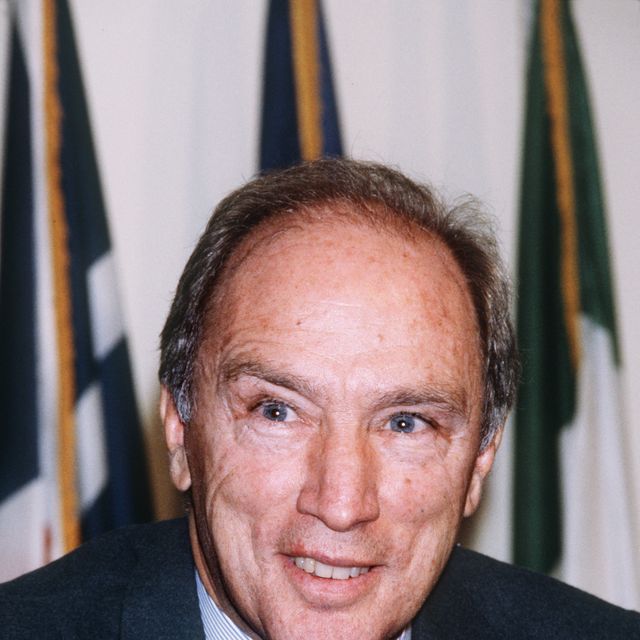 pierre trudeau smiles and looks past the camera, he wears a blue suit jacket, white and blue striped shirt, and dark striped die with a red rose on his lapel