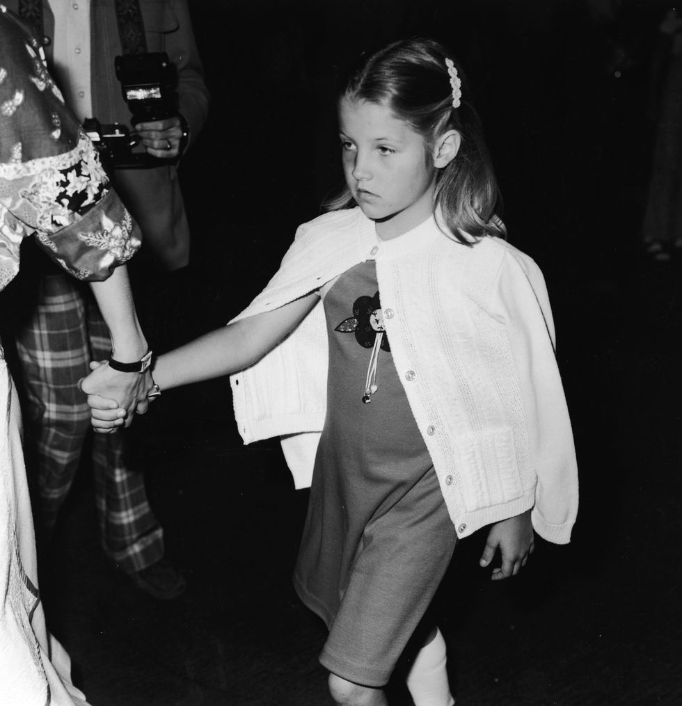 lisa marie presley as a child walks and holds the hand of an adult out of frame, she wears a dress and sweater on her shoulders and buttoned at the collar