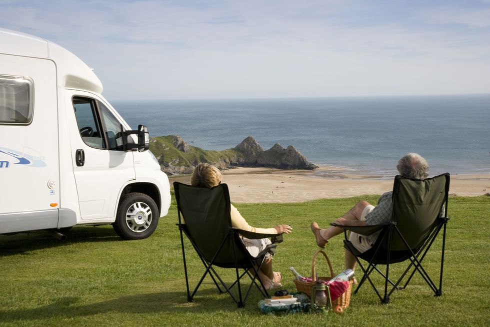 A couple with a picnic relaxing in their chairs next to their motorhome enjoying the view across Three Cliffs Bay on the Gower peninsula in Wales., Three Cliffs Bay, Swansea, Wales.