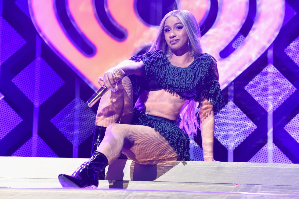 cardi b sits on a step on a stage, she holds a wireless microphone in one hand and wears a two piece outfit with fringe and boots