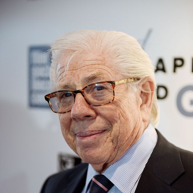carl bernstein smiles at the camera, he is wearing a black suit jacket, blue and white striped collared shirt, a black tie with pink strips, and tortoise shell glasses