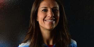 carli lloyd smiles at the camera with a soccer ball resting on top of her head, her blue and white jersey is just visible