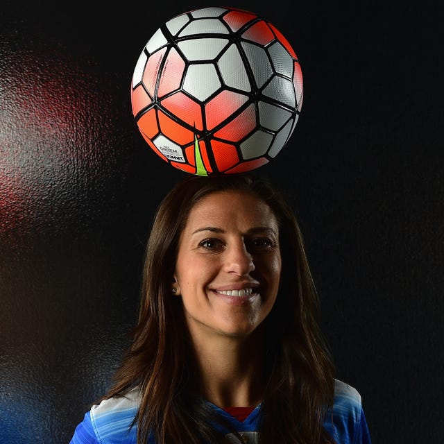 carli lloyd smiles at the camera with a soccer ball resting on top of her head, her blue and white jersey is just visible