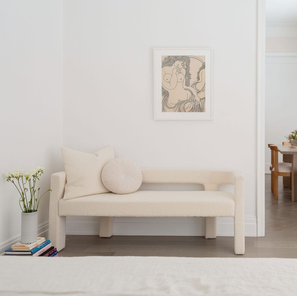 very white room with a long upholstered bench with cutouts along the wall  with a small table and wooden chairs in the far room behind it