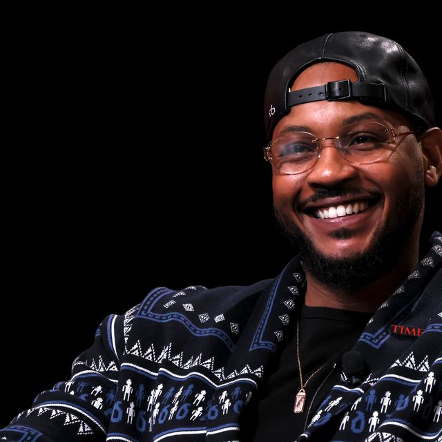 carmelo anthony smiles and looks past the camera, he is wearing glasses, a backwards black baseball cap, a black shirt, a black patterned sweater and a silver necklace