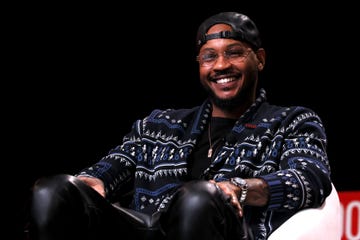 carmelo anthony smiles and looks past the camera, he is wearing glasses, a backwards black baseball cap, a black shirt, a black patterned sweater and a silver necklace