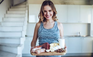 young sporty woman is standing on light kitchen at home with healhy food full of protein in hand, smiling and looking at camera healthy food concept