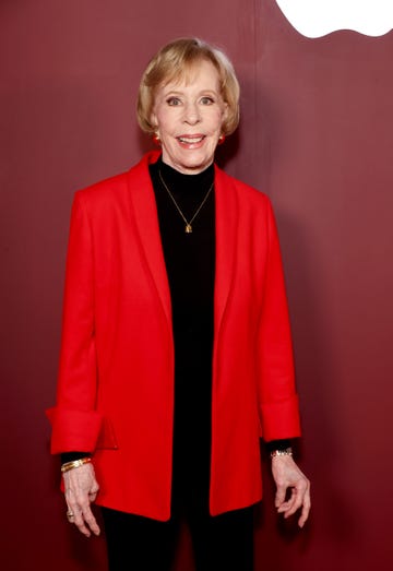 carol burnett smiles at the camera, she wears a bright red jacket over a black shirt with a gold necklace and red earrings