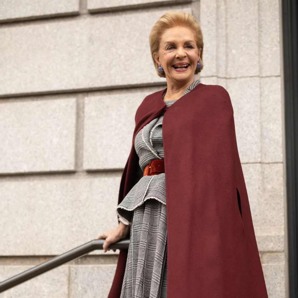 carolina herrera stands outside a stone building and holds a handrail, she smiles and looks right of the camera, she wears a gray plaid skirt suit and a burgundy cape with blue earrings