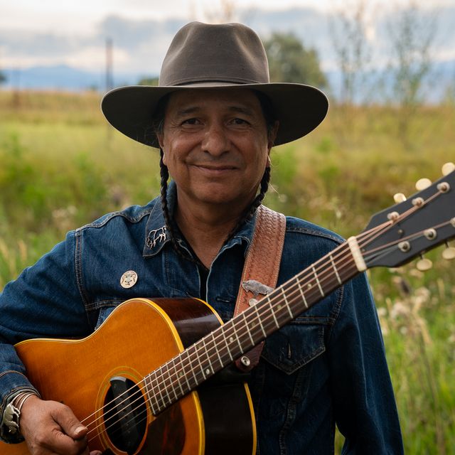 portrait of cary morin, acoustic blues artist holding a guitar in a field, wearing acowboy hat and denim shirt
