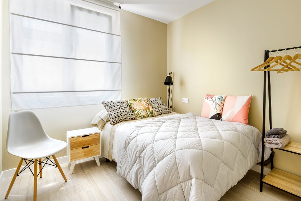 dormitorio decorado en tonos neutros con cojines florales, silla nórdica, mesilla de madera y pequeño vestidor abierto