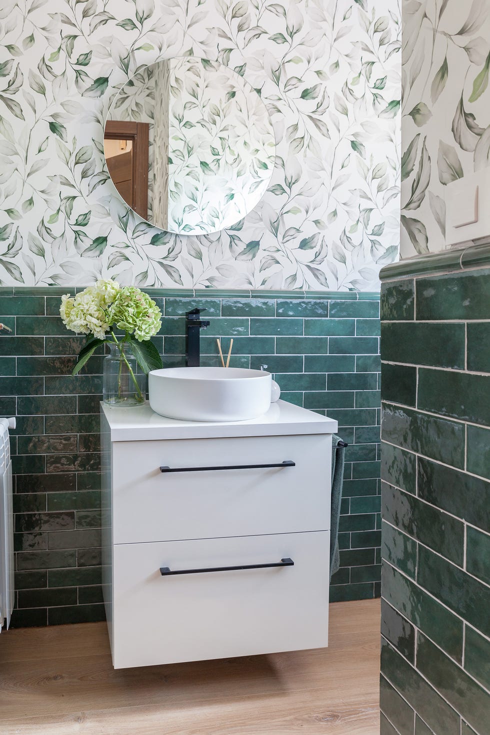 baño con azulejos tipo metro verdes, papel pintado con motivos botánicos y mueble volado blanco