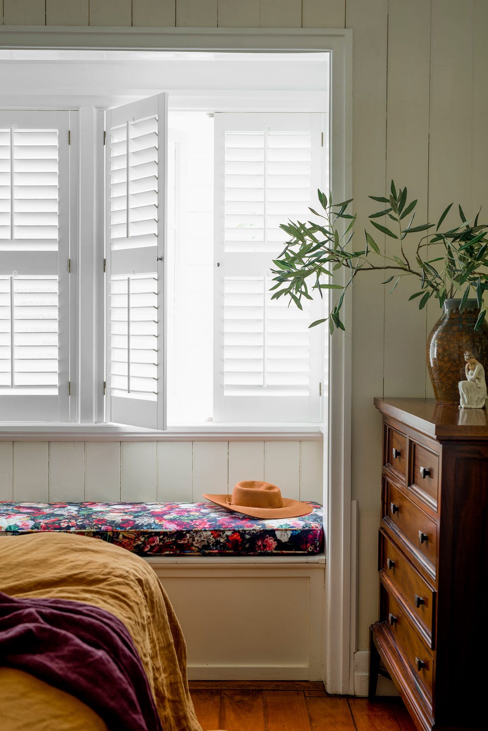 dormitorio de estilo rústico con lamas de madera blanca en las paredes y banco de obra con colchón floral bajo la ventana