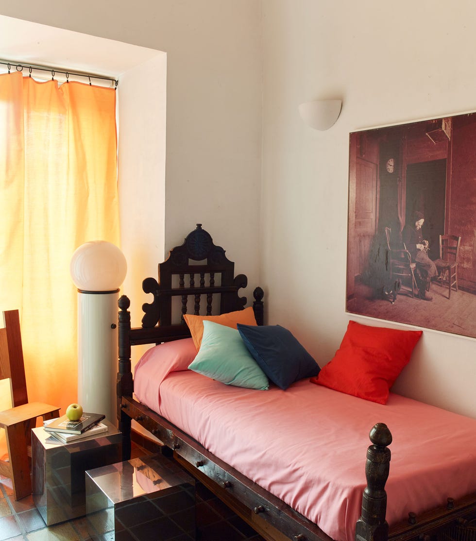 twin bed with wood frame posts and salmon sheets and several pillows and a yellow curtain in the window next to it and a painting over the inside wall of the bed and heavy cubes on the other side as tables