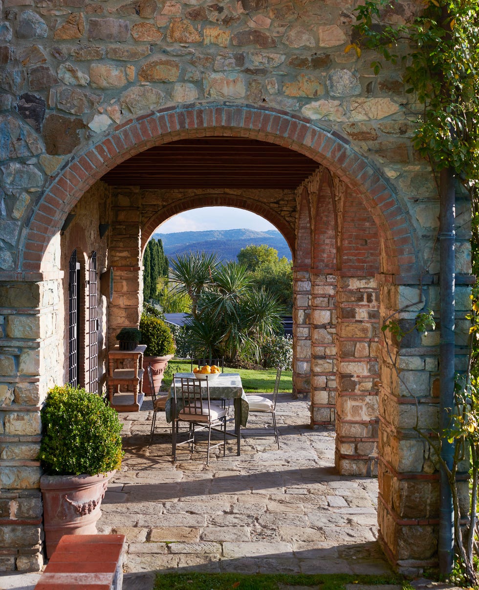straight view through an arched loggia with table and chairs on stone looking out over the green lush tuscan countryside