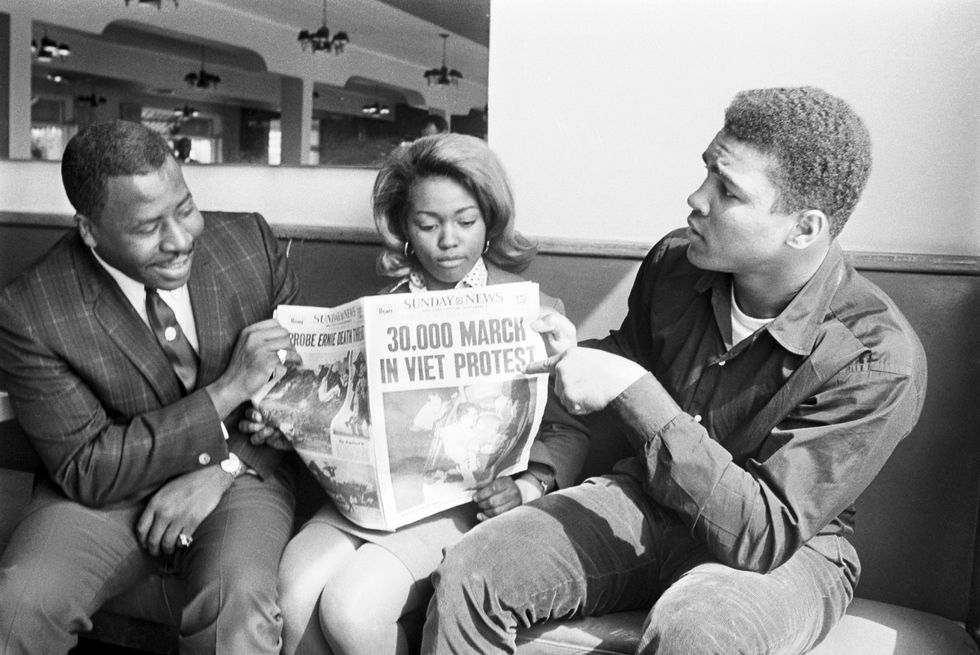 muhammad ali, who sits on a couch, points to a 1966 newspaper headline about a vietnam war protest, the newspaper is held by a man and a woman on ali's left who are also sitting on the couch