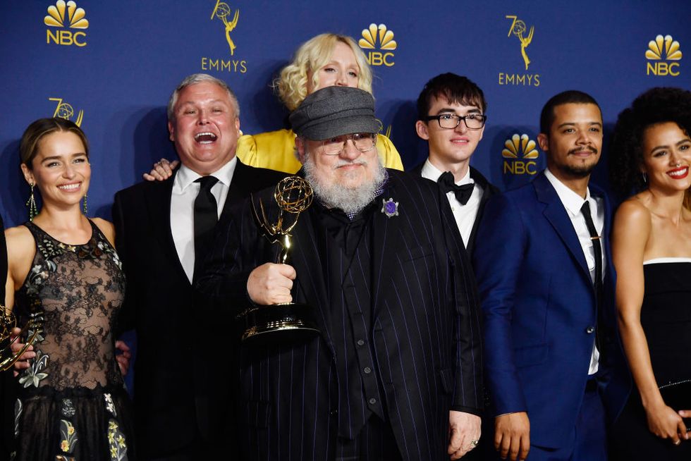 george r r martin standing in front of the game of thrones cast and holding up an emmy trophy