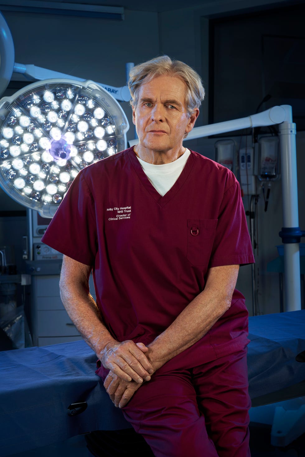 casualty's russell whitelaw sits on the edge of an operating table as a large surgical light shines behind him