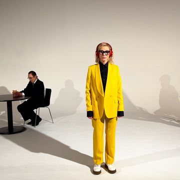cate blanchett wears a yellow suit, black glasses, and red headphones while staring directly into the camera, with ron and russell mael sit in chairs behind her in an all white room
