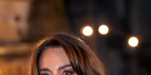 kate middleton smiles and looks left of the camera, she wears a white jacket over a white sweater with dangling earrings, she stands outside with blurred lights in the background