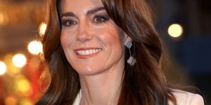 kate middleton smiles and looks left of the camera, she wears a white jacket over a white sweater with dangling earrings, she stands outside with blurred lights in the background