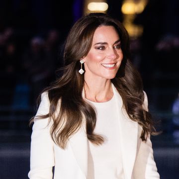 kate middleton walks outdoors and smiles while looking right, she wears a white shirt and coat with white dangling earrings