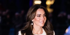 kate middleton walks outdoors and smiles while looking right, she wears a white shirt and coat with white dangling earrings