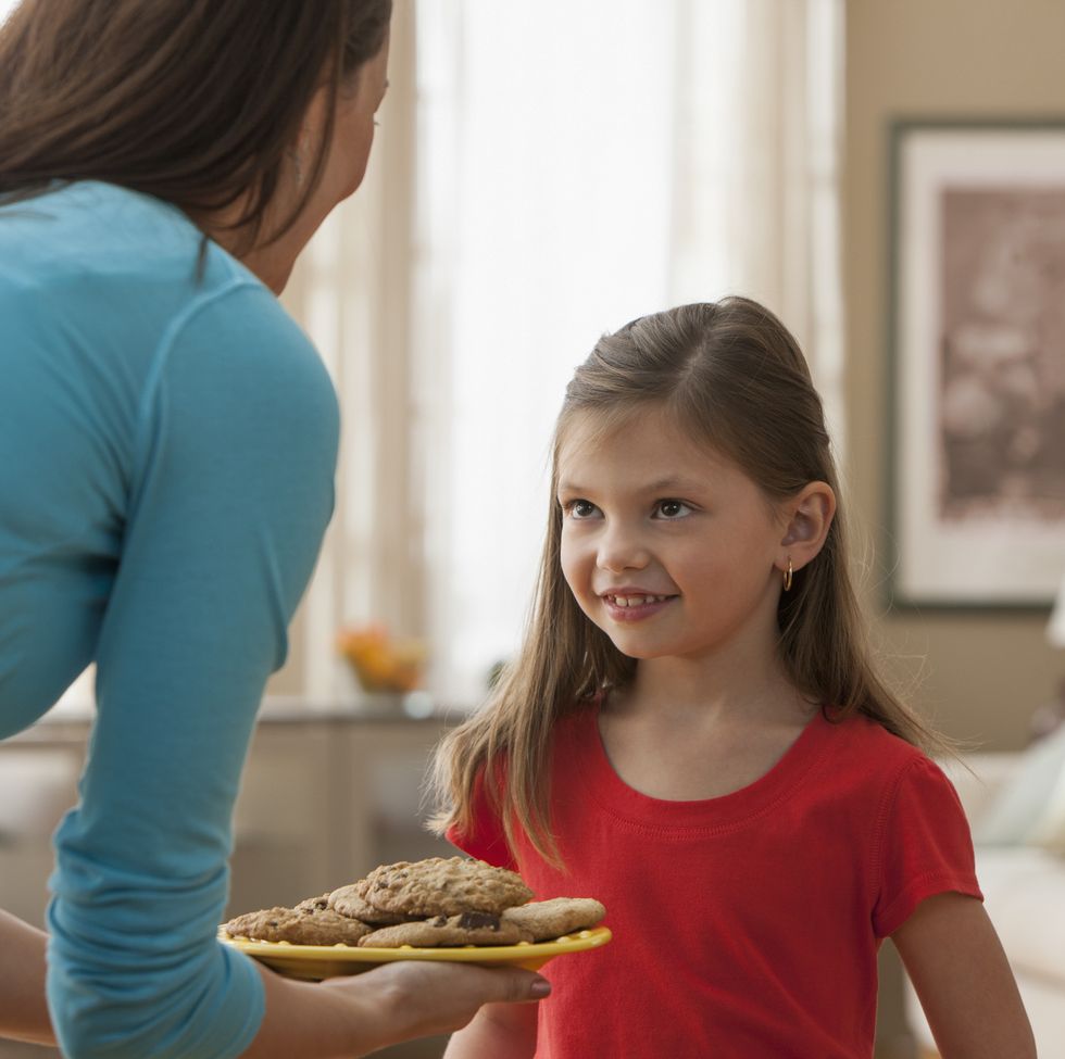 a white mom in a blue shirt offering her daughter in a red shirt a plate of chocolate chip cookies