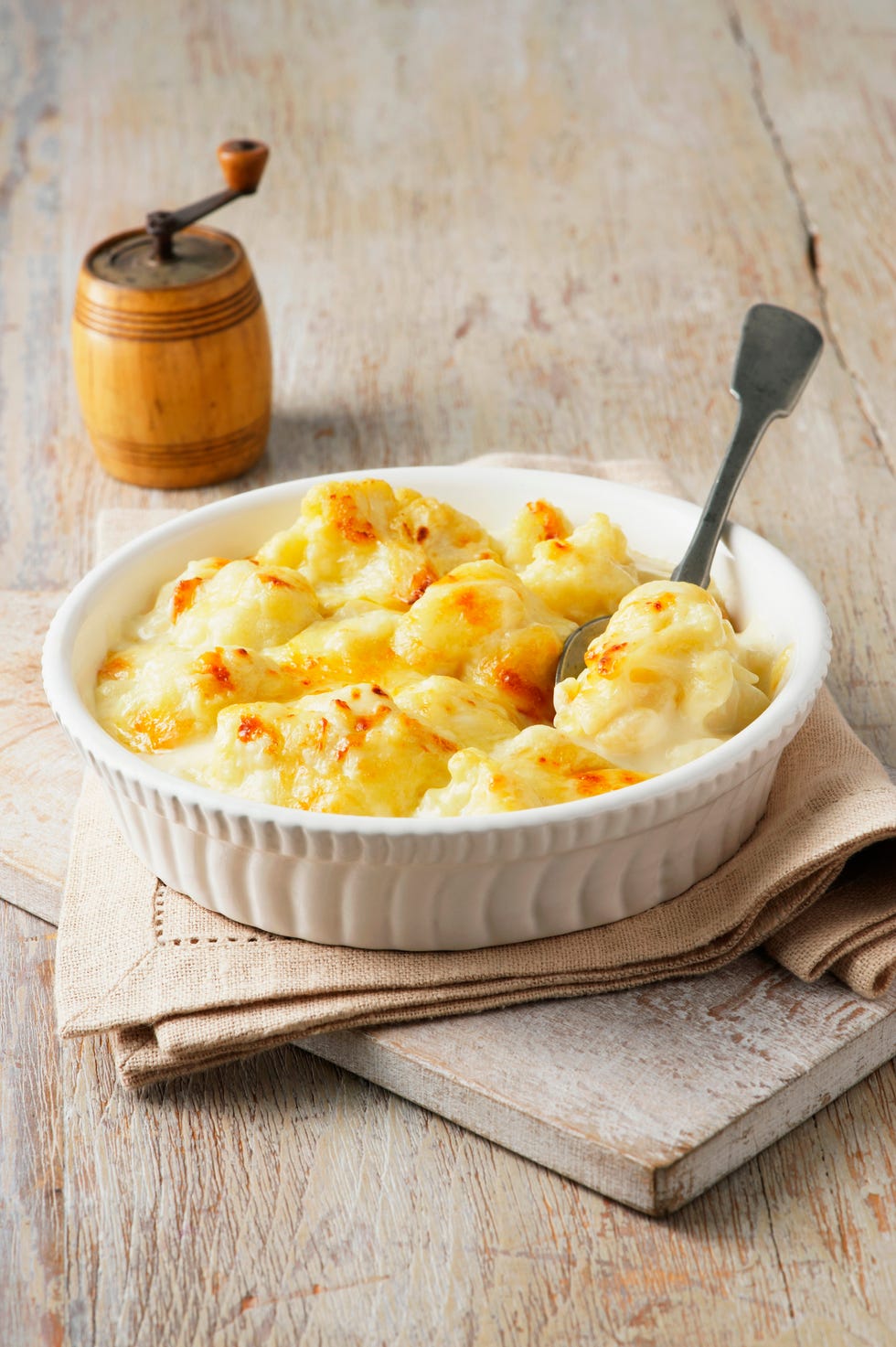 Cauliflower cheese in large bowl with spoon and wooden pepper grinder