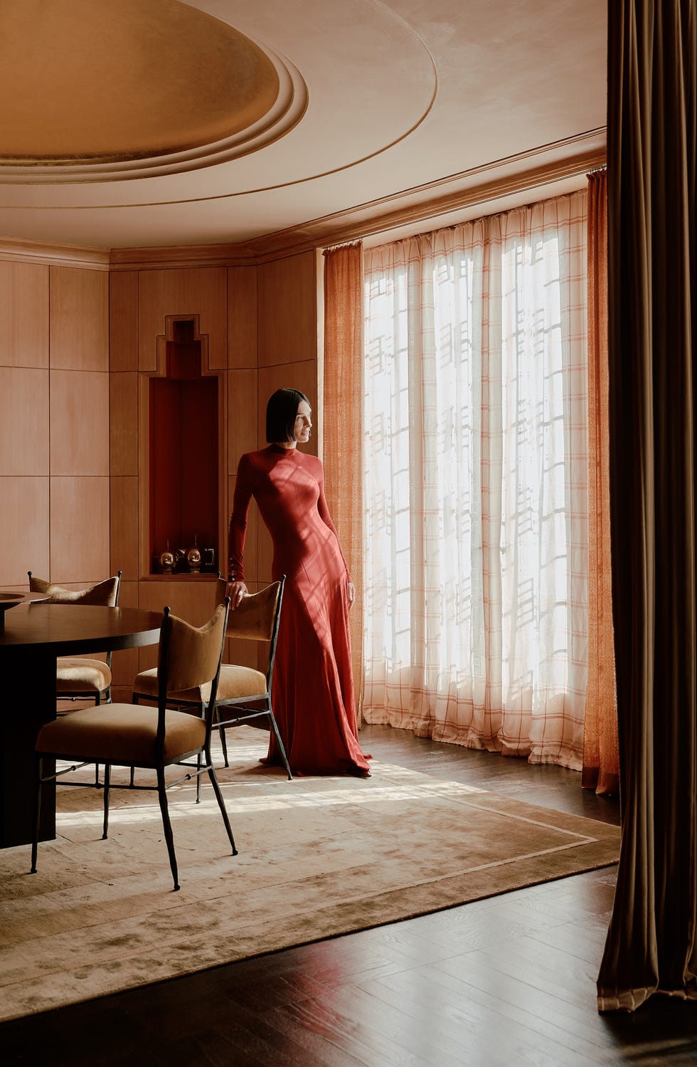 Athena Calderone posant dans une pièce de style art déco, regardant par la fenêtre tout en portant une robe rouge à manches longues