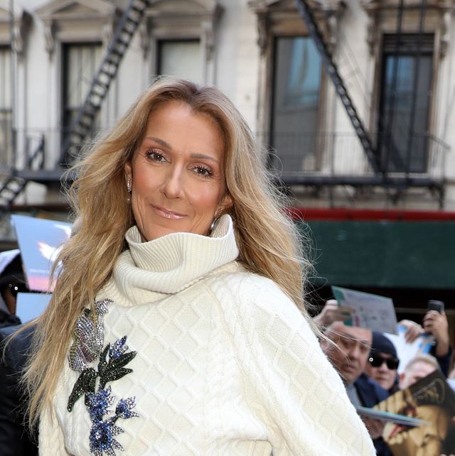 celine dion stands outside and smiles at the camera, she is wearing a white sweater with a larger ribbed collar and blue floral embrodiery
